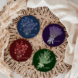 May include: Set of four round coasters featuring botanical illustrations. Designs include blue berries with "les bleuets", lavender with "la lavande", red berries with "les canneberges", and eucalyptus with "eucalyptus". Each coaster has a white ribbon.