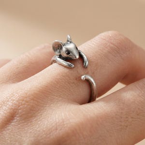 May include: A silver-colored ring featuring a detailed mouse design. The mouse has a textured body, large ears, and small black eyes. The ring wraps around the finger, with the mouse's arms appearing to hug the finger.
