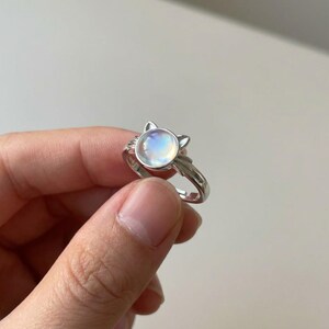 Può includere: Anello color argento con design a forma di gatto. L'anello presenta una pietra rotonda e iridescente per il volto del gatto, con piccole orecchie. La fascia regolabile ha un design semplice ed elegante. La pietra misura circa 1,5 cm di diametro.