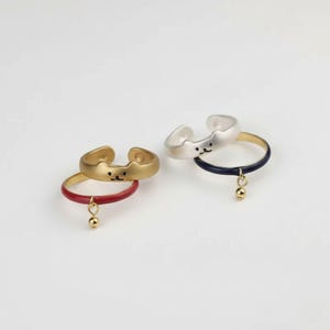 Puede incluir: Cuatro anillos ajustables con diseño de cara de gato. Dos son dorados, uno con una banda roja y un dije dorado, el otro con una banda dorada. Los otros dos son plateados, uno con una banda azul marino y un dije dorado, el otro con una banda dorada.