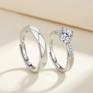 Op de afbeelding: Twee zilveren ringen. De ene ring heeft een geometrisch ontwerp, de andere heeft een grote, heldere edelsteen en kleinere stenen op de band. Beide ringen zijn gemarkeerd met "S925".
