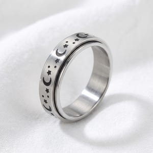 Op de afbeelding: Een zilverkleurige spinnerring met een hemels ontwerp. De ring heeft een band met geëtste halve manen en sterren. De binnenste band draait vrij. De ring is gemaakt van een glanzend, metalen materiaal.