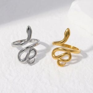 May include: Two snake-shaped rings, one silver and one gold, are shown against a white backdrop. The rings have a coiled design with a raised snake's head. The rings are likely made of metal and are designed to be worn as jewellery.