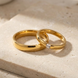 Op de afbeelding: Twee gouden trouwringen. De ene is een eenvoudige band, de andere heeft een kleine, heldere edelsteen. De ringen staan tegen een neutrale, getextureerde achtergrond, wat een klassieke en elegante uitstraling creëert.