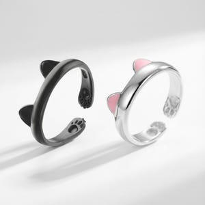 May include: Two adjustable cat-themed rings. One ring is black with black cat ears and paw prints. The other ring is silver with pink cat ears and paw prints. The rings are open-ended.