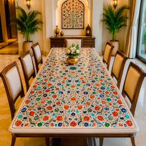 May include: A rectangular dining table with a white stone surface featuring an intricate inlaid floral pattern in red, blue, and green. The table has a dark wood base and is surrounded by six chairs with light-coloured upholstered seats and backs.