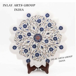 Könnte beinhalten: Eine dekorative Platte aus weißem Marmor mit floralem Intarsienmuster. Die Platte hat einen gewellten Rand und ist mit blauen Blumenmustern und einem zentralen Sternmuster verziert. Der Text "INLAY ARTS GROUP INDIA" ist sichtbar. Die Platte wird auf einem Holzständer präsentiert.
