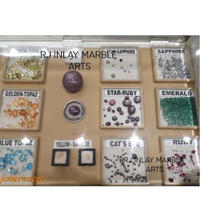 K&ouml;nnte beinhalten: Eine Vitrine mit verschiedenen Edelsteinen, darunter Goldtopas, Blautopas, Smaragd und Rubin. Die Vitrine zeigt auch Sternsaphir-, Sternrubin- und Katzenaugensteine. Der Text "RJ Inlay Marble Arts" ist sichtbar.