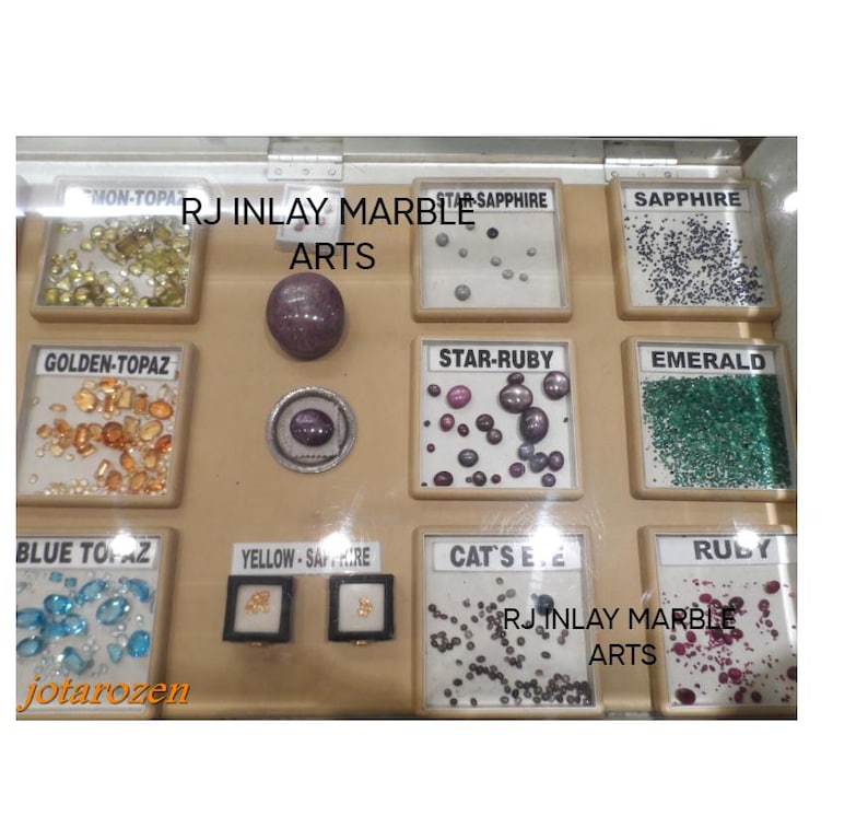 K&ouml;nnte beinhalten: Eine Vitrine mit verschiedenen Edelsteinen, darunter Goldtopas, Blautopas, Smaragd und Rubin. Die Vitrine zeigt auch Sternsaphir-, Sternrubin- und Katzenaugensteine. Der Text "RJ Inlay Marble Arts" ist sichtbar.