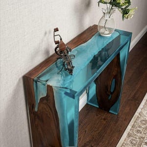 Peut inclure: Une console moderne avec un cadre en bois et un dessus en résine époxy turquoise. La table présente un design à bord brut et une sculpture en métal. Le texte "Epoxy Treasures House" est visible.