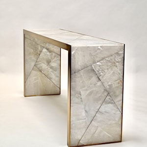 Pode incluir: Uma consola moderna com um design geométrico. A mesa apresenta uma superfície cinza claro, semelhante a pedra, com um padrão de formas irregulares. As bordas são emolduradas com metal dourado. A mesa tem aproximadamente 122 cm de comprimento.