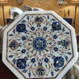 Könnte beinhalten: Achteckiger weißer Marmortisch mit eingelegtem Blumendesign. Das Design zeigt blaue und goldene Blumen und Blätter mit einem zentralen Sternmuster. Der Tisch hat einen braunen Rand und den Text "INLAY ARTS GROUP INDIA".