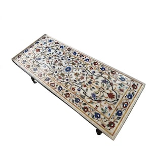 May include: Rectangular white marble table with intricate floral inlay design in shades of blue, red, and green. The table has a dark border and four short legs. The overall design is ornate and detailed.