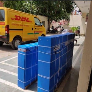 K&ouml;nnte beinhalten: Ein gelber DHL-Lieferwagen steht neben mehreren gro&szlig;en, rechteckigen Paketen, die in blaues Papier gewickelt und mit wei&szlig;em Klebeband befestigt sind. Die Pakete sind mit "HANDLE IN BLUE" beschriftet.