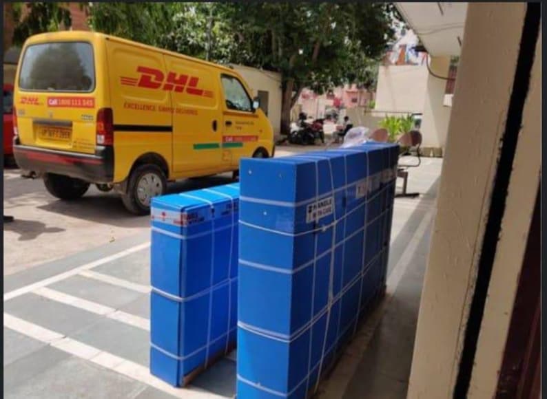 K&ouml;nnte beinhalten: Ein gelber DHL-Lieferwagen steht neben mehreren gro&szlig;en, rechteckigen Paketen, die in blaues Papier gewickelt und mit wei&szlig;em Klebeband befestigt sind. Die Pakete sind mit "HANDLE IN BLUE" beschriftet.