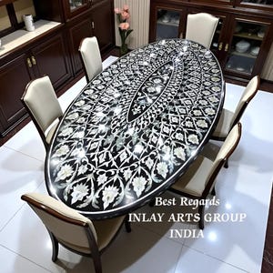 Puede incluir: Una mesa de comedor ovalada negra con un diseño floral incrustado en blanco y plata. Seis sillas color crema rodean la mesa. El texto "Best Regards INLAY ARTS GROUP INDIA" es visible.