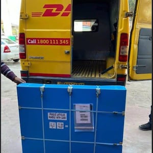 K&ouml;nnte beinhalten: Ein blauer rechteckiger Versandbeh&auml;lter mit Metallverschl&uuml;ssen und einem wei&szlig;en Etikett wird in einen gelben DHL-Lieferwagen geladen. Der Beh&auml;lter tr&auml;gt die Aufschrift "FRAGILE ARTICLES HANDLE WITH CARE". Der Transporter hat das DHL-Logo und eine Telefonnummer.