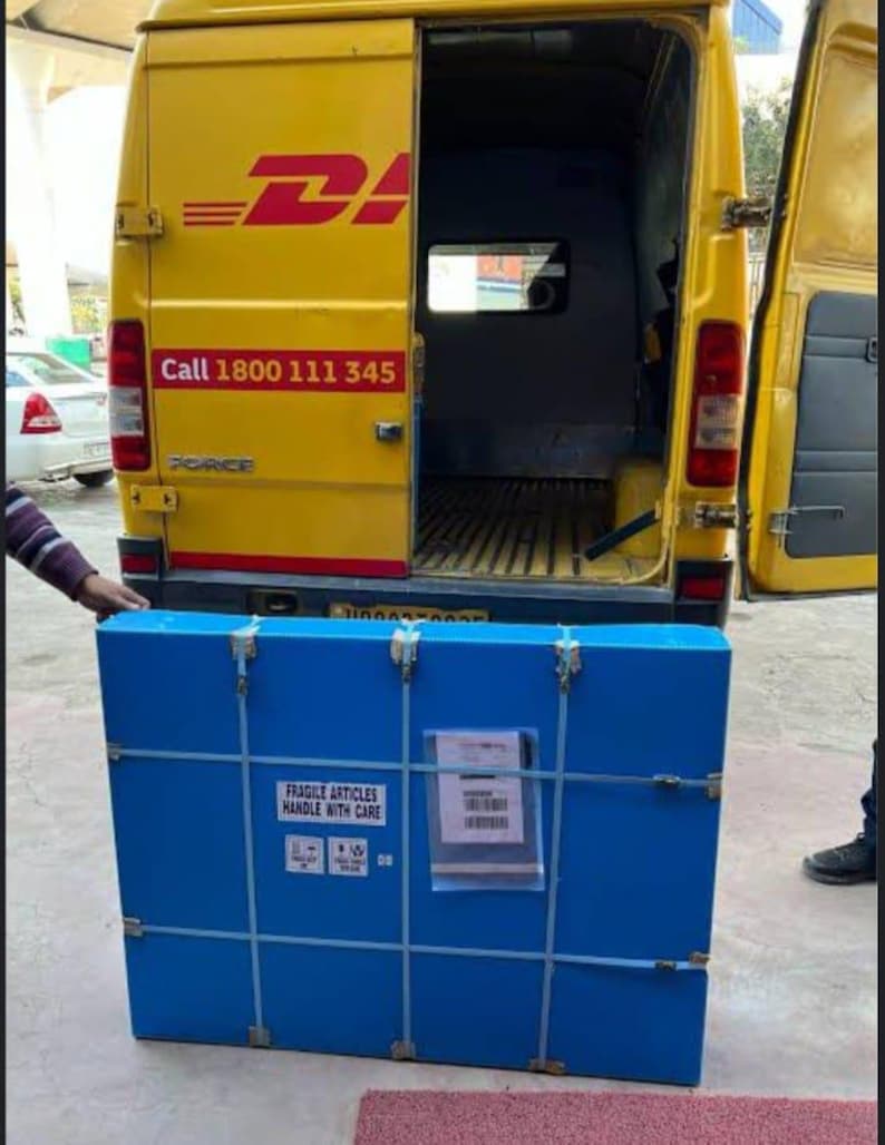 K&ouml;nnte beinhalten: Ein blauer rechteckiger Versandbeh&auml;lter mit Metallverschl&uuml;ssen und einem wei&szlig;en Etikett wird in einen gelben DHL-Lieferwagen geladen. Der Beh&auml;lter tr&auml;gt die Aufschrift "FRAGILE ARTICLES HANDLE WITH CARE". Der Transporter hat das DHL-Logo und eine Telefonnummer.
