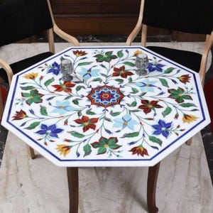 Pode incluir: Mesa octogonal de mármore branco com um design floral embutido. A mesa apresenta uma borda azul e é suportada por quatro pernas marrons. O padrão floral inclui flores vermelhas, azuis, verdes e amarelas. Dois objetos decorativos prateados estão sobre a mesa.
