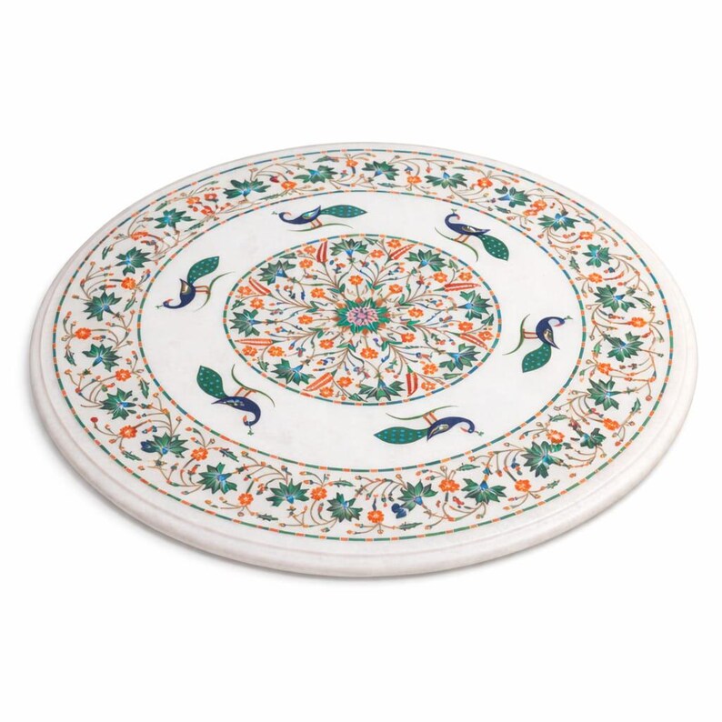 K&ouml;nnte beinhalten: Eine runde Tischplatte aus wei&szlig;em Marmor mit einem aufwendigen Intarsienmuster. Das Design zeigt ein zentrales Blumenmedaillon, Pfauen und florale Muster in Gr&uuml;n, Orange und Blau. Der Durchmesser der Tischplatte betr&auml;gt ca. 91 cm.