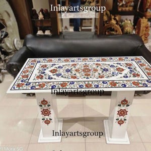 Op de afbeelding: Witte marmeren tafel met een rechthoekig blad en bloemeninlegontwerp in rood, blauw en oranje. De tafel heeft een witte basis met decoratieve bloempatronen. De tekst "Inlayartsgroup" is zichtbaar.