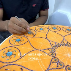 K&ouml;nnte beinhalten: Ein Handwerker arbeitet an einer orangefarbenen Marmoroberfl&auml;che und platziert sorgf&auml;ltig blaue und t&uuml;rkisfarbene Einlegearbeiten. Das Design zeigt komplizierte Blumenmuster und einen Rand aus kleinen blauen Steinen. Der Text "INLAY ARTS GROUP INDIA" ist sichtbar.