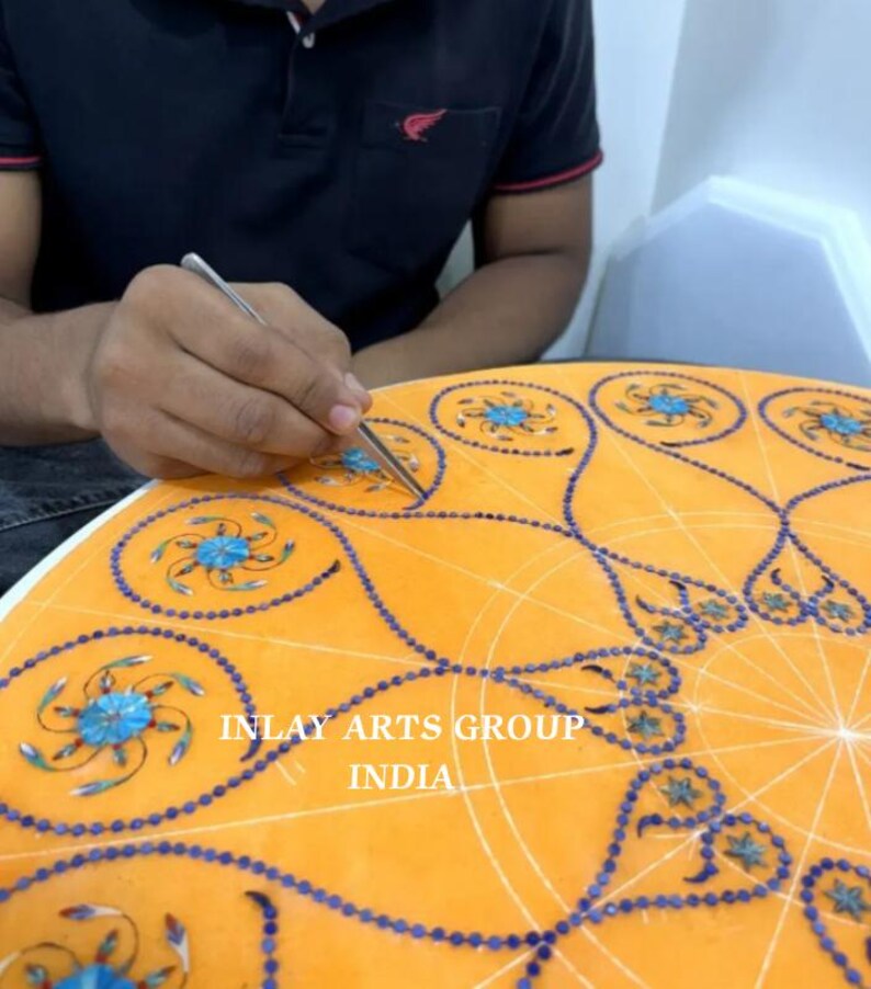 K&ouml;nnte beinhalten: Ein Handwerker arbeitet an einer orangefarbenen Marmoroberfl&auml;che und platziert sorgf&auml;ltig blaue und t&uuml;rkisfarbene Einlegearbeiten. Das Design zeigt komplizierte Blumenmuster und einen Rand aus kleinen blauen Steinen. Der Text "INLAY ARTS GROUP INDIA" ist sichtbar.