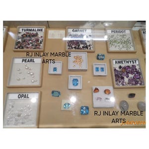 K&ouml;nnte beinhalten: Eine Ausstellung verschiedener Edelsteine, darunter Turmalin, Granat, Peridot, Perle, Opal, Amethyst und Topas. Jeder Edelstein ist beschriftet und in einer kleinen, quadratischen Vitrine pr&auml;sentiert. Der Text "RJ INLAY MARBLE ARTS" ist sichtbar.