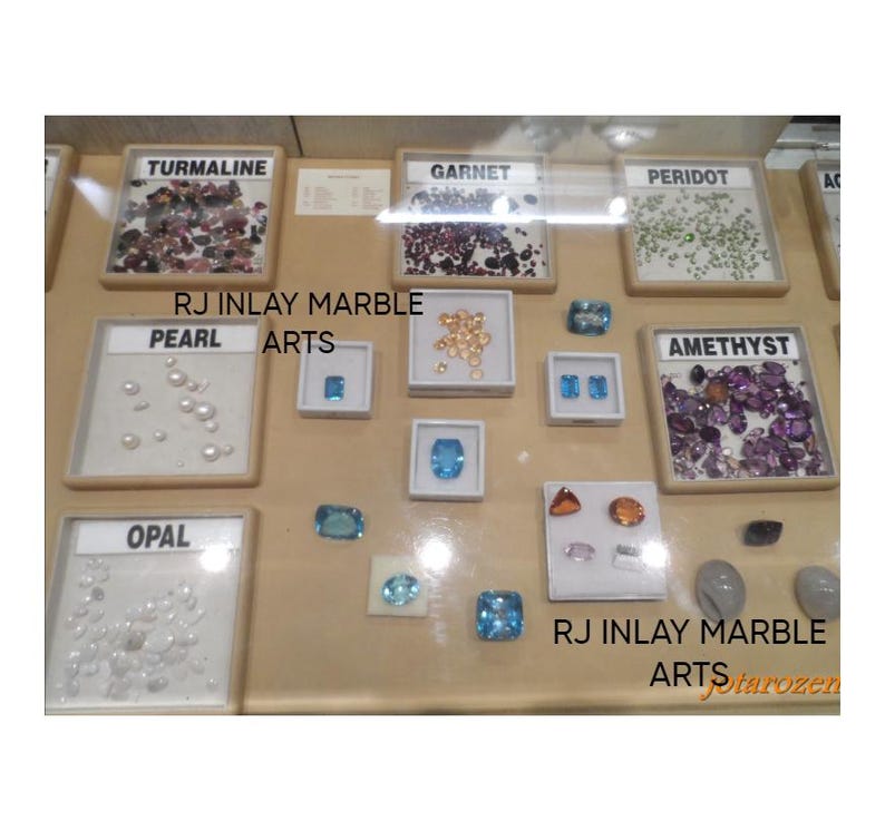 K&ouml;nnte beinhalten: Eine Ausstellung verschiedener Edelsteine, darunter Turmalin, Granat, Peridot, Perle, Opal, Amethyst und Topas. Jeder Edelstein ist beschriftet und in einer kleinen, quadratischen Vitrine pr&auml;sentiert. Der Text "RJ INLAY MARBLE ARTS" ist sichtbar.