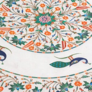 K&ouml;nnte beinhalten: Wei&szlig;er Marmor-Tischplatte mit aufwendigem Blumen- und Pfauendesign. Das Design weist orange, gr&uuml;ne, blaue und rosa Akzente auf. Die Tischplatte hat eine kreisf&ouml;rmige Form und dient wahrscheinlich dekorativen Zwecken.