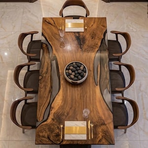 Op de afbeelding: Een bovenaanzicht van een eettafel met acht houten stoelen. De tafel heeft een live edge design met een donkere houtnerf en een heldere harsinleg. Een kom met decoratieve stenen staat in het midden. Plaatsinstellingen en een kroonluchter zijn ook zichtbaar.