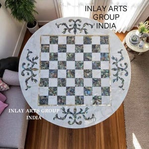 Puede incluir: Una mesa de ajedrez redonda de mármol blanco con una superficie de juego a cuadros. La mesa tiene detalles florales decorativos y el texto "INLAY ARTS GROUP INDIA". Los cuadrados del tablero de ajedrez son blancos e iridiscentes.