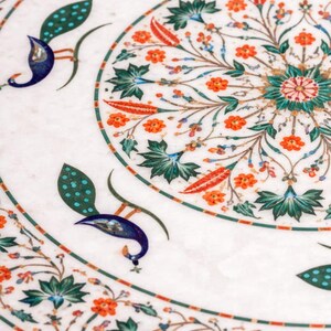 K&ouml;nnte beinhalten: Nahaufnahme einer wei&szlig;en Marmoroberfl&auml;che mit aufwendigen Blumen- und Pfauenfedermotiven. Das Design zeigt orangefarbene Blumen, gr&uuml;ne Bl&auml;tter und blaue Pfauenelemente. Die Details sind vor einem wei&szlig;en Hintergrund.