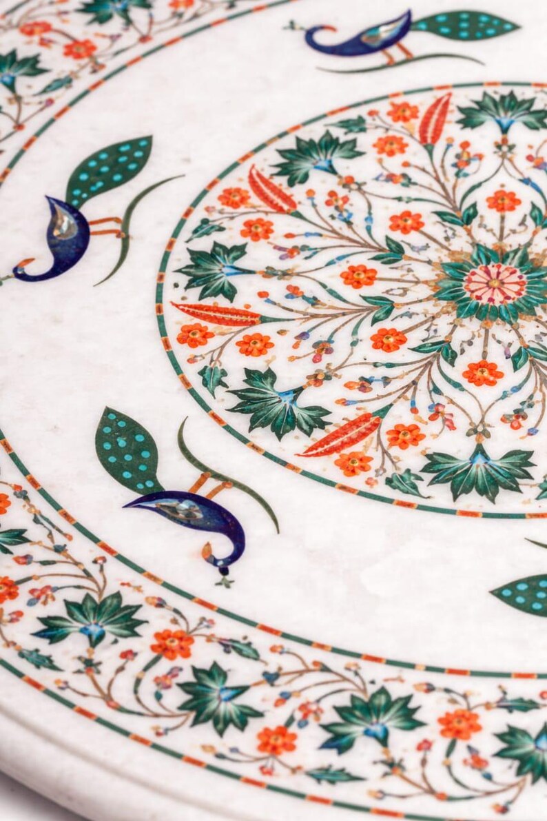 K&ouml;nnte beinhalten: Nahaufnahme einer wei&szlig;en Marmoroberfl&auml;che mit aufwendigen Blumen- und Pfauenfedermotiven. Das Design zeigt orangefarbene Blumen, gr&uuml;ne Bl&auml;tter und blaue Pfauenelemente. Die Details sind vor einem wei&szlig;en Hintergrund.
