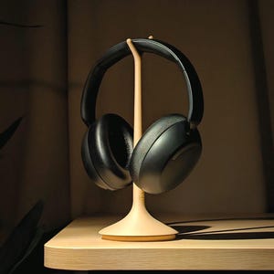 May include: Black over-ear headphones on a light wood headphone stand. The stand has a flared base and a slender, vertical support. The headphones are set against a neutral background, ideal for showcasing audio accessories.