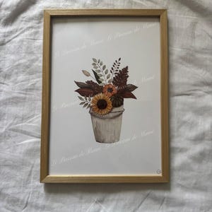 Peut inclure: Une impression encadrée présentant une illustration à l'aquarelle d'un bouquet de tournesols, de feuilles séchées et de feuillage dans un seau rustique. Le texte Le Pinceau de Mamé est répété sur l'image. L'œuvre est dans un cadre en bois clair.