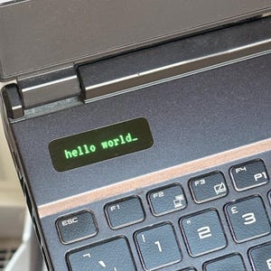 Op de afbeelding: Een donkergrijs laptoptoetsenbord met een klein, rechthoekig digitaal scherm ingebed boven de toetsen. Het scherm toont de groene tekst "hello world_" in een monospaced lettertype. Het toetsenbord bevat standaard functietoetsen.
