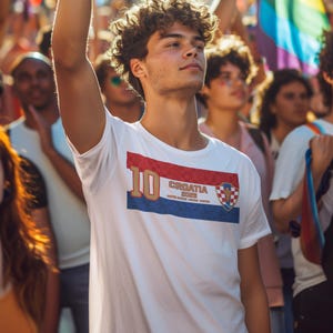 May include: White t-shirt with a Croatian flag design. The shirt features a red, white, and blue rectangular graphic with the number "10" in gold, the word "CROATIA" and a shield emblem. The shirt is worn by a person with curly hair.