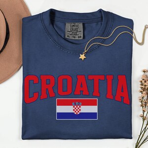 May include: Navy blue t-shirt featuring the word "CROATIA" in red and the Croatian flag. A gold necklace with a star pendant is on the shirt. The shirt has a Comfort Colors label.