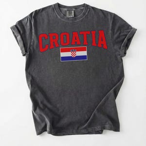 May include: Dark grey t-shirt featuring the word "CROATIA" in red, arched lettering. Beneath the text is the Croatian flag. The shirt has rolled-up sleeves and a crew neck.