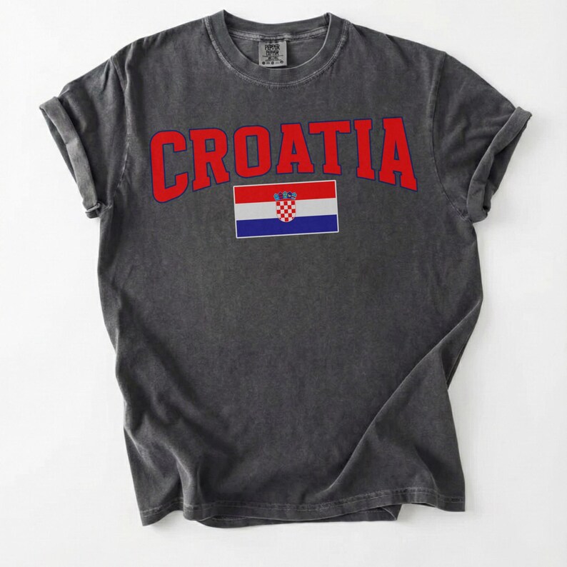 May include: Dark grey t-shirt featuring the word "CROATIA" in red, arched lettering. Beneath the text is the Croatian flag. The shirt has rolled-up sleeves and a crew neck.