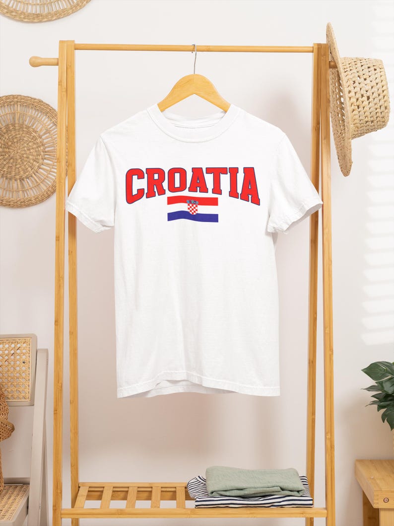 May include: White t-shirt with the word "CROATIA" in red block letters and the Croatian flag underneath. The shirt is hanging on a wooden clothes rack with a straw hat.