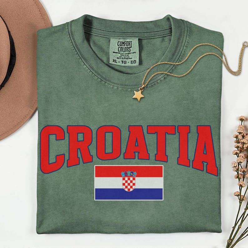 May include: A green t-shirt featuring the word "CROATIA" in red lettering and a Croatian flag. The shirt has a crew neck and short sleeves. A gold necklace with a star pendant is on the shirt.