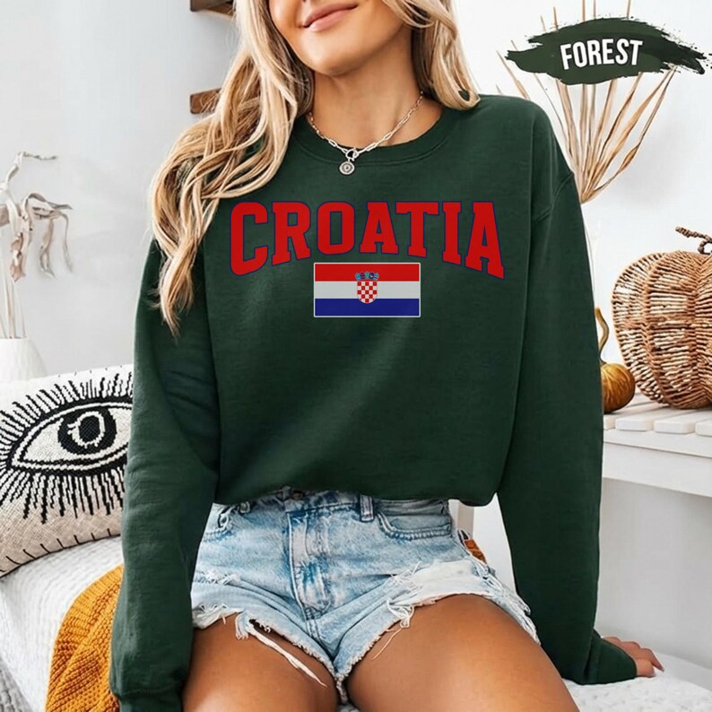 May include: A dark green sweatshirt featuring the word "CROATIA" in red above the Croatian flag. The flag is a horizontal tricolour of red, white, and blue with the Croatian coat of arms in the centre. The sweatshirt has a crew neck.