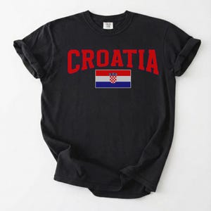 May include: Black t-shirt featuring the word "CROATIA" in red, arched text. Beneath the text is the Croatian flag. The shirt is made of a soft material and has rolled-up sleeves.