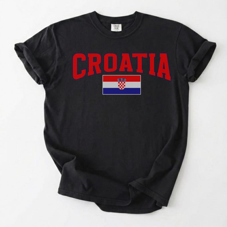 May include: Black t-shirt featuring the word "CROATIA" in red, arched text. Beneath the text is the Croatian flag. The shirt is made of a soft material and has rolled-up sleeves.