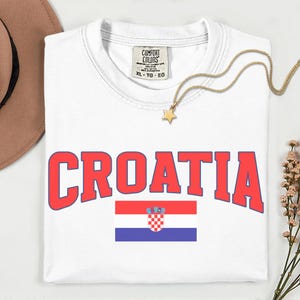 May include: White t-shirt featuring the word "CROATIA" in red block letters with a blue outline. The Croatian flag is below the word. A gold necklace with a star pendant lies on the shirt. The shirt has a Comfort Colors tag.