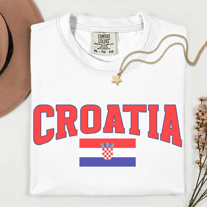 May include: White t-shirt featuring the word "CROATIA" in red block letters with a blue outline. The Croatian flag is below the word. A gold necklace with a star pendant lies on the shirt. The shirt has a Comfort Colors tag.