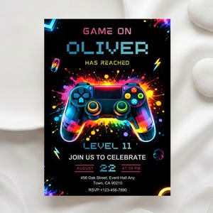 May include: A black invitation card with a colorful video game controller graphic. The text reads "GAME ON OLIVER HAS REACHED LEVEL 11 JOIN US TO CELEBRATE" with event details. The design features vibrant splashes of color.