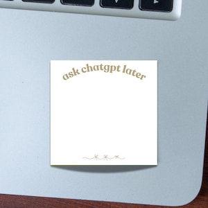 以下が含まれることがあります： 「ask chatgpt later」というテキストが薄茶色で書かれた白い付箋。 付箋の下部には小さなリボンの装飾線があります。 付箋はシルバーのノートパソコンの上に置かれています。